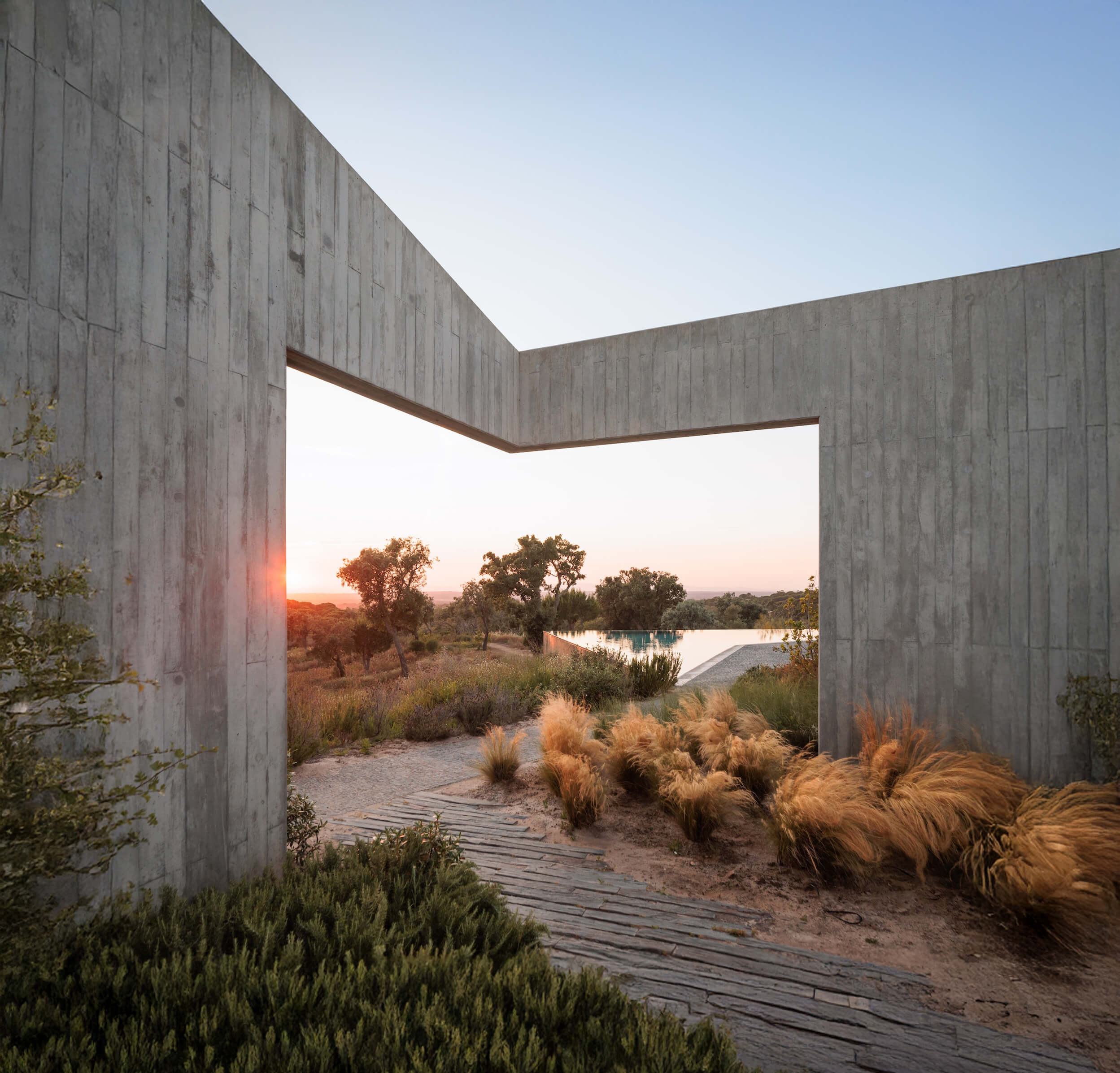 A large, modern concrete wall with a rectangular opening frames a scenic sunset view at a two bedroom villa in Portugal, with sunlight streaming through trees and tall grasses in a natural landscape.
