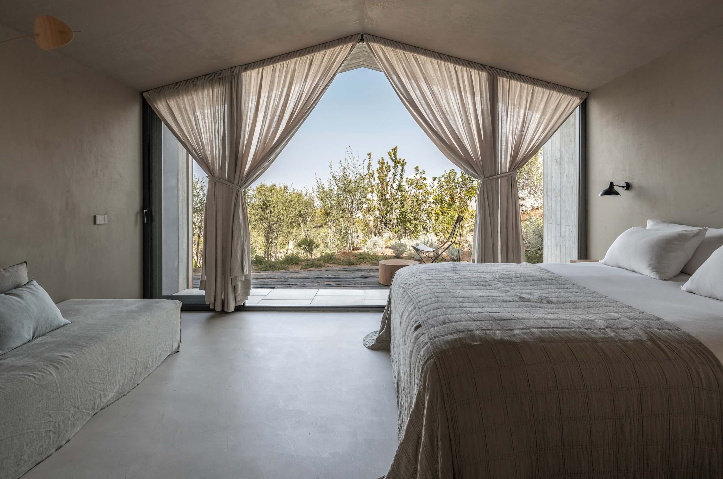 A serene bedroom in a hotel in Portugal with light gray walls, a large bed draped in a textured quilt, a sofa, and floor-to-ceiling windows with sheer curtains overlooking a lush outdoor garden and stone patio.