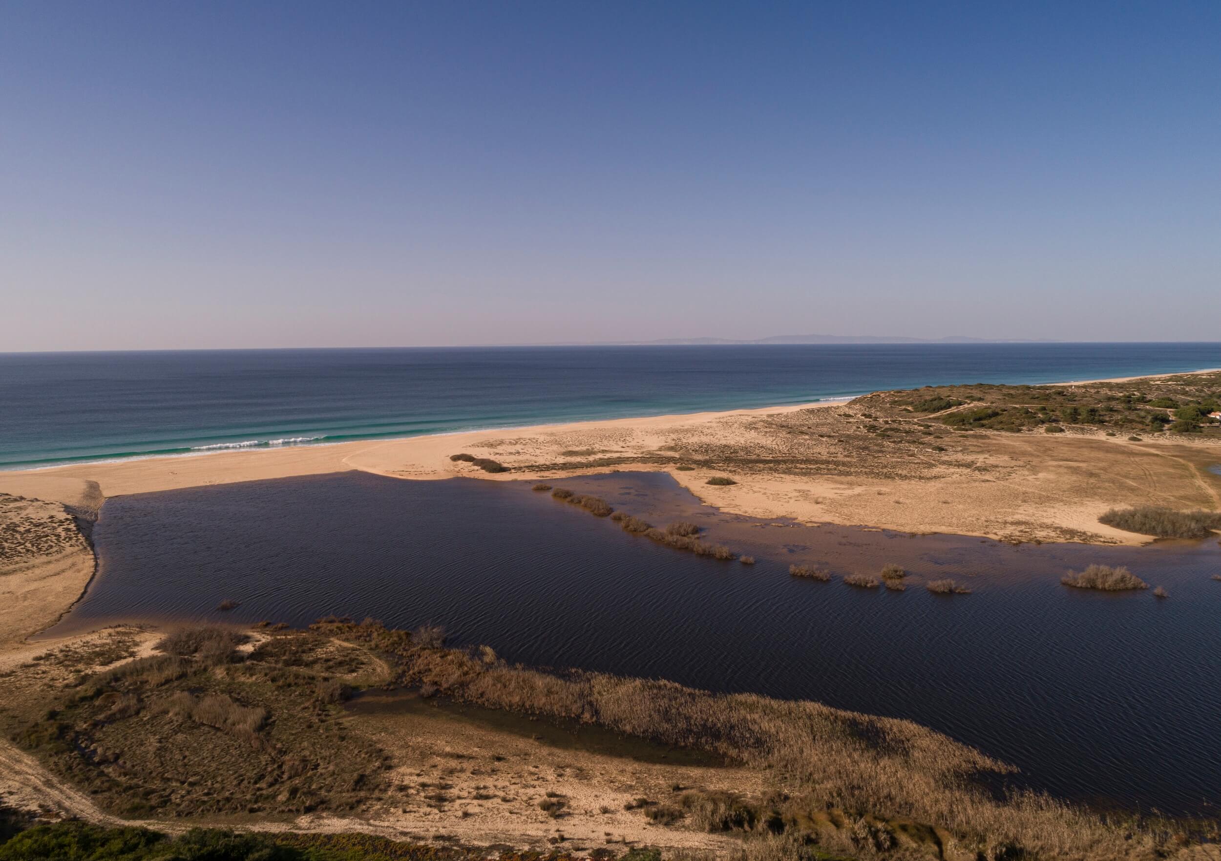 Aerial view of a sandy beach along the coast of Alentejo with a calm lagoon in the foreground, dunes in the middle, and the blue ocean stretching to the horizon under a clear sky. Sparse vegetation surrounds the lagoon and dunes.