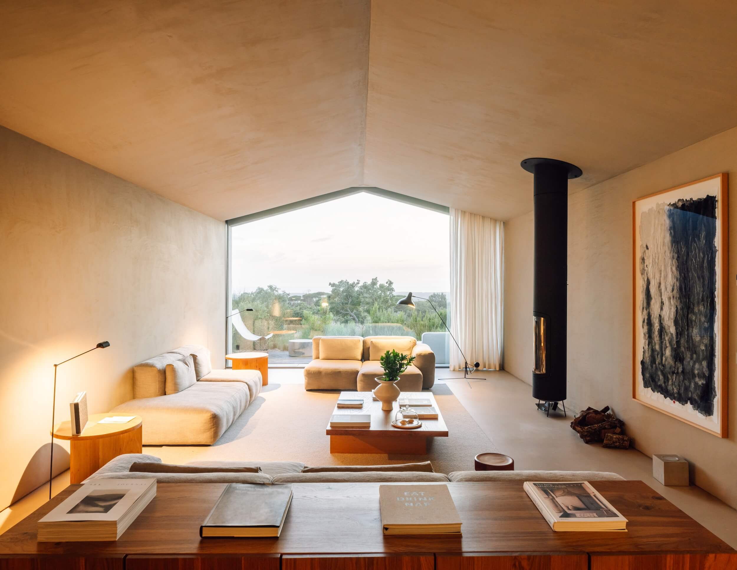 A modern living room in Portugal with neutral tones, large beige sofas, a wood stove, and a floor-to-ceiling window overlooking lush greenery. Minimalist decor includes books, artwork, and abundant natural light filling the space.