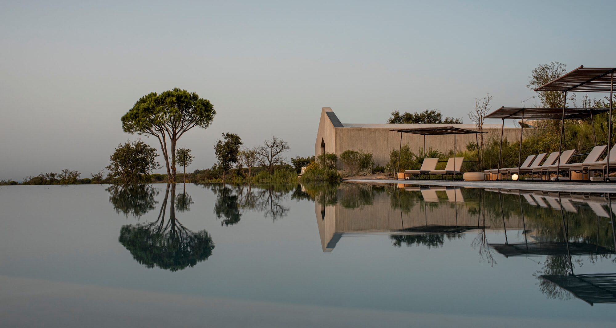 An infinity pool at this hotel in Portugal reflects nearby trees, lounge chairs, and a modern, minimalist building under a clear sky, creating a tranquil and serene atmosphere.