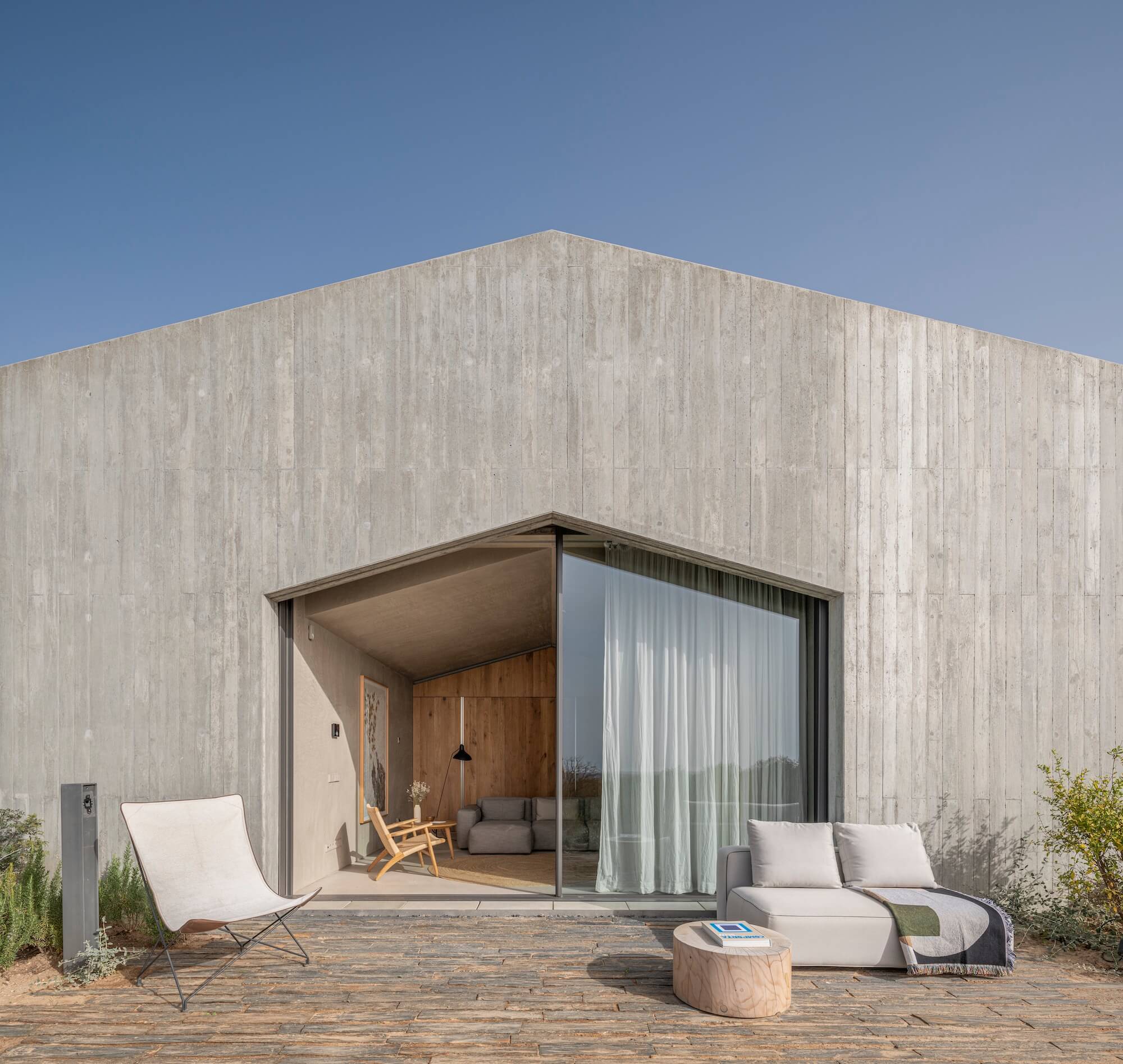A modern house with a minimalist concrete facade echoes the style of luxury villas in Portugal, featuring a geometric entrance, sliding glass doors, light outdoor furniture, and a wooden patio against a clear blue sky.
